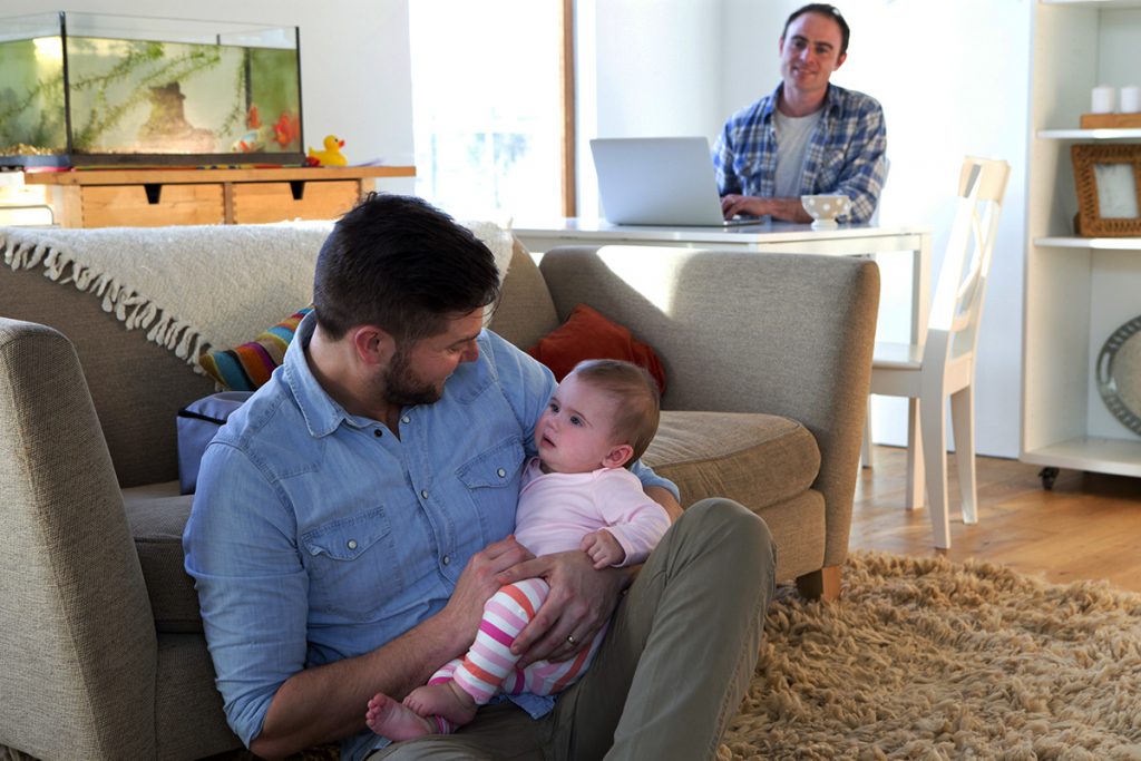 Same sex male couple sitting in their home. One father is sitting on the floor with their daughter in his arms.