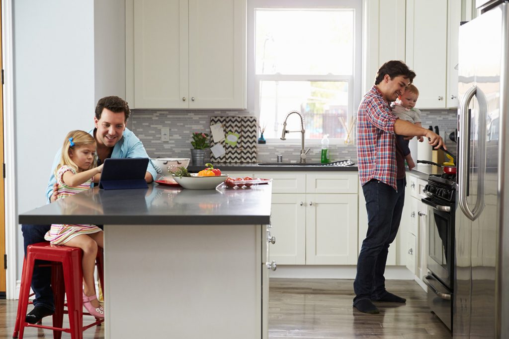 Girl uses tablet in kitchen with dad, while other dad cooks