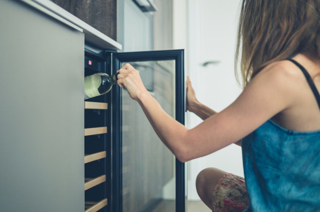 A young woman is getting a bottle of white wine from her wine cooler at home