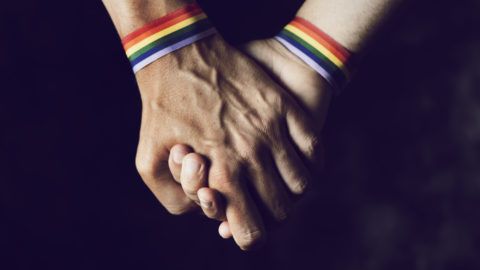 closeup of two caucasian men holding hands with a rainbow-patterned wristban on their wrists
